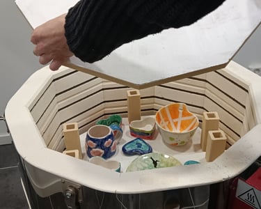 A hand opening a ceramic kiln filled with colorful glazed pottery bowls and mugs.