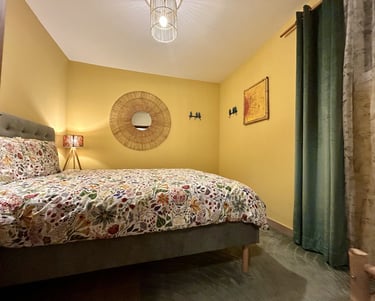 Cozy bedroom with yellow walls, floral bedding, a wicker mirror, and soft ambient lighting.