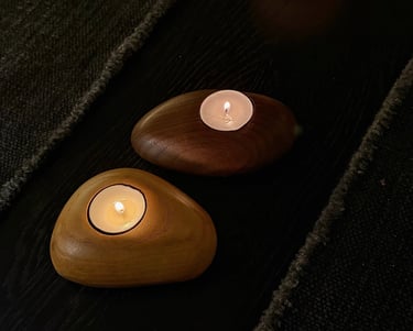 Two lighted tea light candles in smooth, pebble-shaped wooden holders on a dark surface.