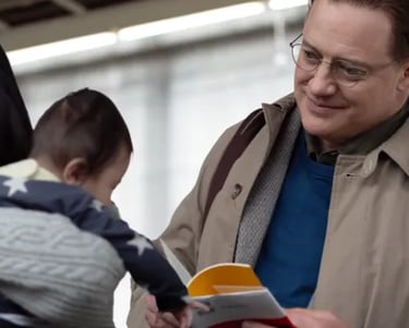 Brendan Fraser smiling gently while interacting with a young child in a warm, everyday setting — pro