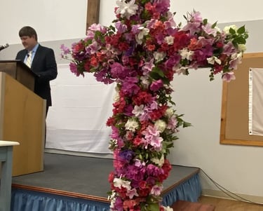 Pastor Wesley Daniels in the pulpit speaking and a large cross covered in Easter flowers