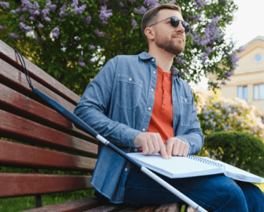 Man with diabetic retinopathy sitting on park bench with cane for the blind reading Louis Braille