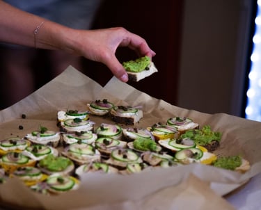 A hand picking up a small piece of bread with avocado on top from a large tray of similar snacks