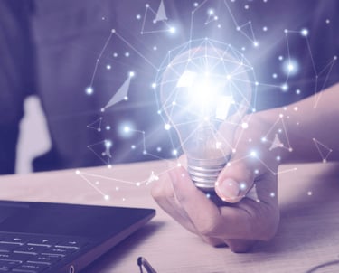 A person's hand holding a light bulb next to a laptop.