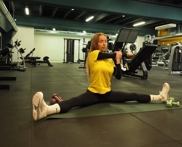 Una donna in maglia gialla esegue uno stretching in spaccata seduta, allungando il braccio per un esercizio di mobilità,