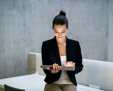 A businesswoman in a black jacket, sitting on a desk, holding a tablet computer.