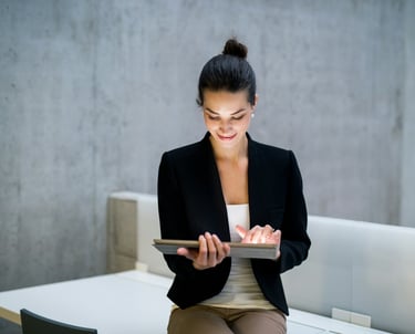 A businesswoman in a black jacket, sitting on a desk, holding a tablet computer.