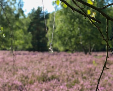 HeideSchmuck Silberkette mit tropfenförmigem Kristallanhänger, aufgehängt an einem Ast vor blühendem Heidekraut in der Natur.
