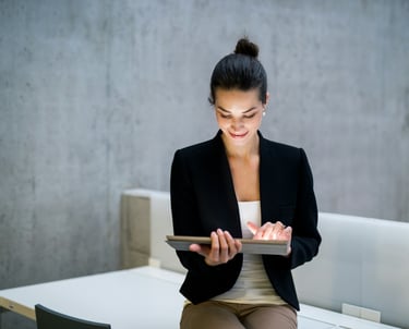 A businesswoman in a black jacket, sitting on a desk, holding a tablet computer.