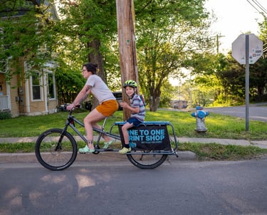 A women riding a bike with a young boy sitting on the back rack with a sign that reads "One to One"