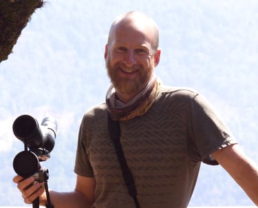 San Cristobal bird guide Benedict Simmons smiling with telescope, with binoculars in front hillside