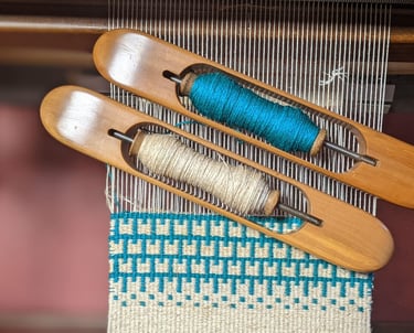Blue and white krokbragd weaving is currently being done on a weaving loom with wooden shuttles and