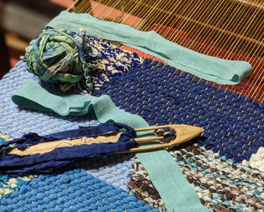 a weaving loom with a wooden shuttle, a ball of rag threads and a rag rug in shades of blue in progr