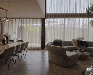 Sala-comedor con cortinas traslúcidas de doble altura, sofá en escuadra y mesa de mármol | KARÄNA