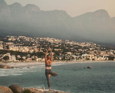 Woman doing yoga on a rocky beach with a coastal city and mountains in the background.