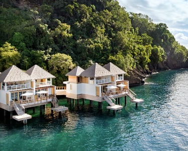 Luxury overwater bungalows at a tropical resort nestled against a lush green cliffside by the ocean.