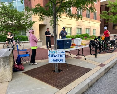 People standing around a table with food and drink behind a sign that reads "Breakfast Station"