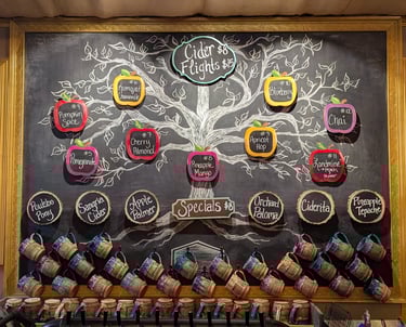 Hand-drawn chalkboard menu at a cidery displaying craft cider flavors, flight prices, and ceramic mugs.
