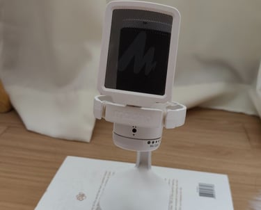 White Maono microphone with pop filter on a stand, placed on a book, with curtain background.