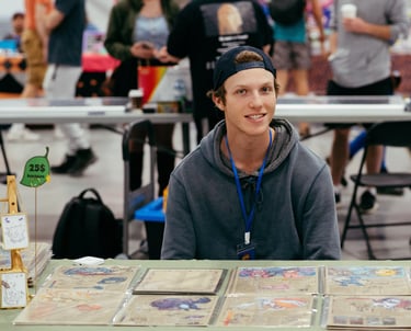 A smiling vendor sits at a table in front of his trading cards for sale.