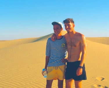  Two young men, smiling and embracing, stand in a desert landscape. The man on the left wears a stri