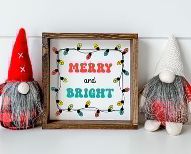 Two Christmas gnomes with a framed print reading “merry and bright” surrounded by colorful Christmas lights