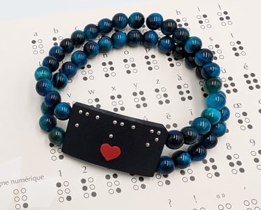 Bracelet perle bleue avec une plaque en braille et un cœur rouge, posé sur un alphabet braille.