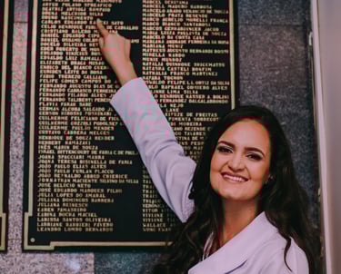 Dra Bruna Melchior de jaleco branco apontando seu nome gravado em placa metálica no hospital das clinicas da USP de ribeirao 