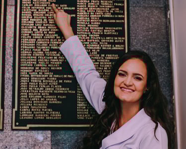 Dra Bruna Melchior de jaleco branco apontando seu nome gravado em placa metálica no hospital das clinicas da USP de ribeirao
