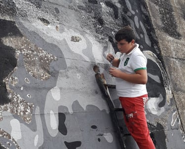Young artist painting a grayscale mural on an outdoor wall, project curated by Poliops.