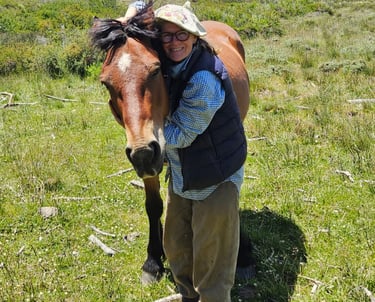 A person hugging a horse in a grassy field with hills in the background.