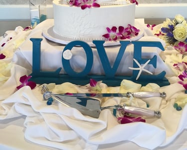 three-tier white wedding cake decorated with fuchsia orchids and a teal“love” sign in front, 