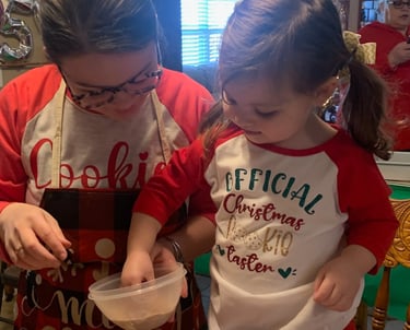 Little girl baking cookies with her aunt wearing a shirt reading, “Official Christmas Cookie Taster”