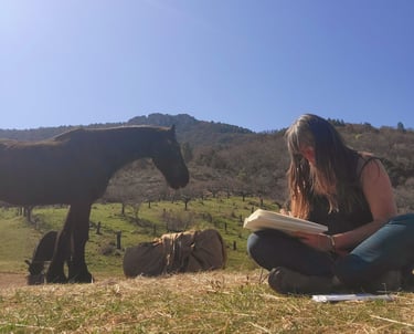 a woman sitting on the grass with a book in her hands and a horse behind her