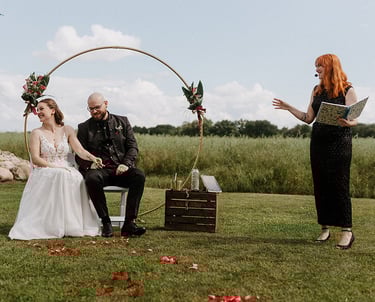 grinsend sitzen Braut und Bräutigam nebeneinander während die Traurednerin im schwarzen Glitzerkleid eine Anekdote erzählt