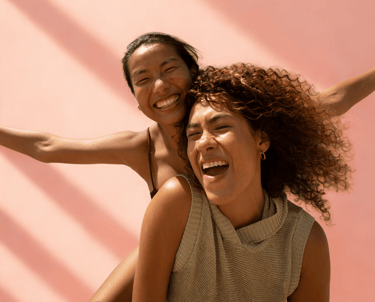 Twee lachende vrouwen, één draagt de ander op de rug tegen een roze achtergrond.