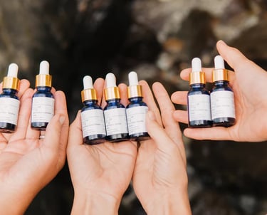 Hands holding blue glass dropper bottles of chakra essential oil blends against a natural stone background.