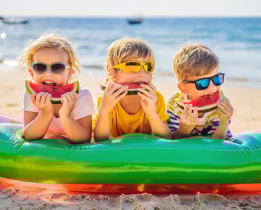 Three young children wearing sunglasses eating fresh watermelon slices on a sunny beach.