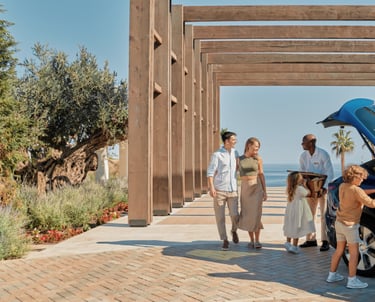 A family arrives at a luxury ikos resort as a hotel porter helps unload luggage from their blue car.