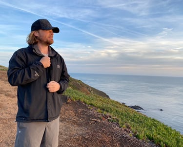 A man in a hiking jacket and cap stands on a coastal cliff overlooking the ocean at sunset.