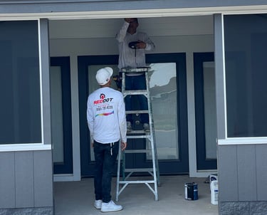 Professional Red Dot painters using a ladder to apply exterior trim paint to a residential porch ceiling.