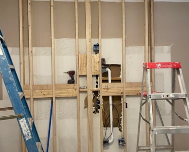 Indoor wall framing renovation with wooden studs, electrical wiring, and plumbing pipes visible between two ladders.