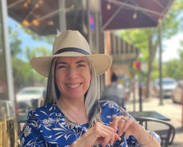 Poet Sheena Powell smiling at an outdoor cafe, wearing a blue patterned shirt and a straw hat
