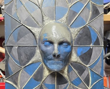 wall hanging relief sculpture in earthy greens, blues and blacks. A face protruding from a geometric web design.