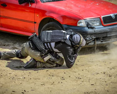A tactical shooter in full gear aims a blue training pistol from behind a white car at a shooting range.