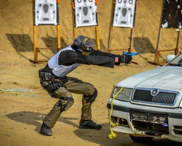 Tactical shooter in training gear aims a blue handgun from behind a car at an outdoor firing range.