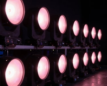 Horizontal shot of a row of stage lights on a North American / US stage, all glowing in a sequence of soft pink hues.