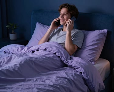A serene North American bedroom at night. A person looks relaxed and happy while talking on a phone, wrapped in a soft blanket. The colors are deep midnight blue and soft lavender, suggesting premium comfort and support.