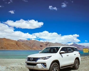 White Toyota Fortuner SUV parked by the scenic Pangong Lake in Ladakh under a blue sky.