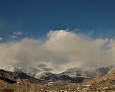Panoramic view of a Himalayan mountain range with snow-capped peaks and scattered clouds over a town in Ladakh.